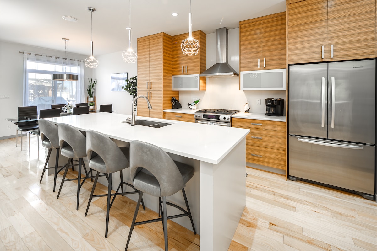 A modern kitchen features light wood cabinetry paired with stainless steel appliances. An oversized island with a sleek countertop is surrounded by high-backed stools, providing ample seating. Natural light streams through a nearby window, illuminating the open and spacious design.