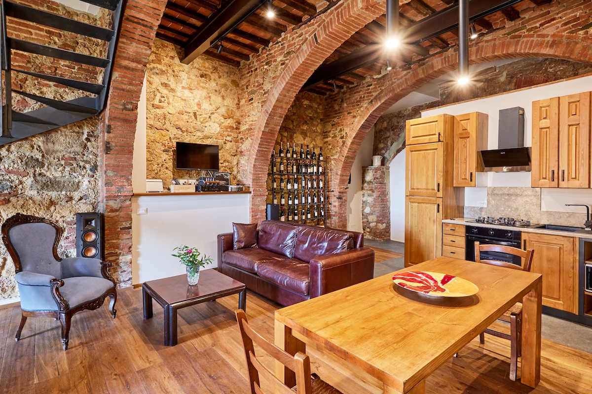 An open-plan living area features exposed stone walls and wooden beams, accentuated by arched architecture. A cozy leather sofa faces a wooden dining table, while a well-equipped kitchen is visible in the background. A decorative wine display adds character to the space.
