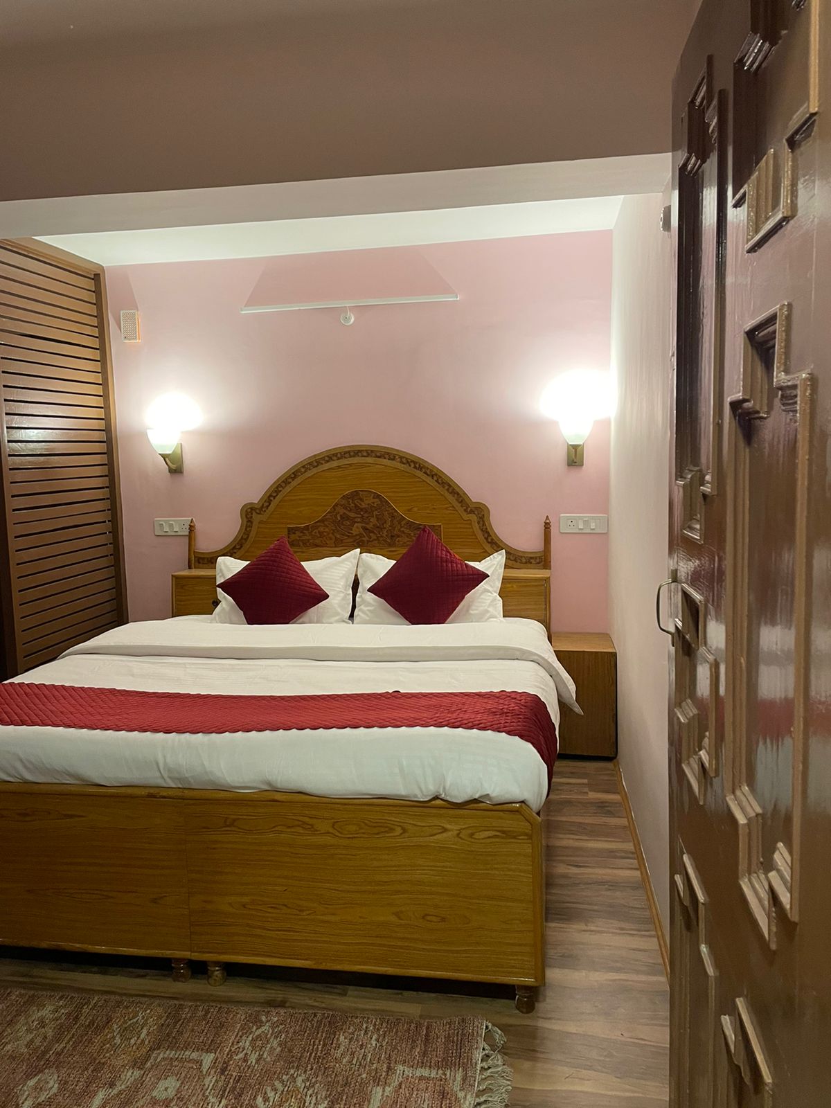 A warm-toned bedroom features a wooden bed with a patterned red blanket and contrasting white linens. Decorative pillows in deep red are placed on the bed. Ambient lighting is provided by wall sconces, and wood paneling adds texture to the space.