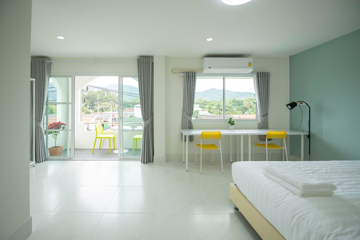 The spacious room includes an expansive kingsize bed, a bright workspace with a table and two yellow chairs, and a large window that opens to a private balcony. Light-colored walls enhance the airy ambiance, complemented by natural light and mountain views in the background.