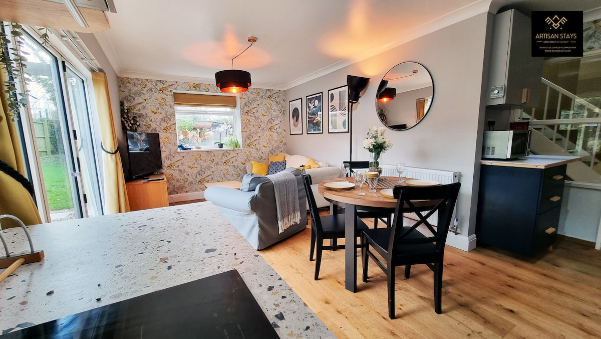 A modern living space is presented, featuring a comfortable gray sofa adorned with vibrant cushions. A round dining table with black chairs is positioned near a window, while artwork decorates the walls. A television is visible, contributing to a cozy atmosphere.