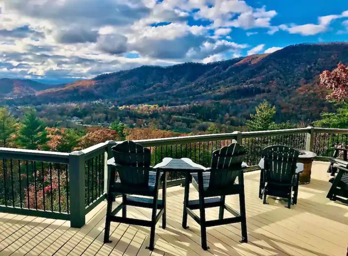 An expansive deck offers panoramic views of the surrounding mountains, showcasing a vibrant autumn landscape. Four black Adirondack chairs surround a small table, inviting relaxation and enjoyment of the scenery. The sky features soft clouds, enhancing the natural beauty of the environment.