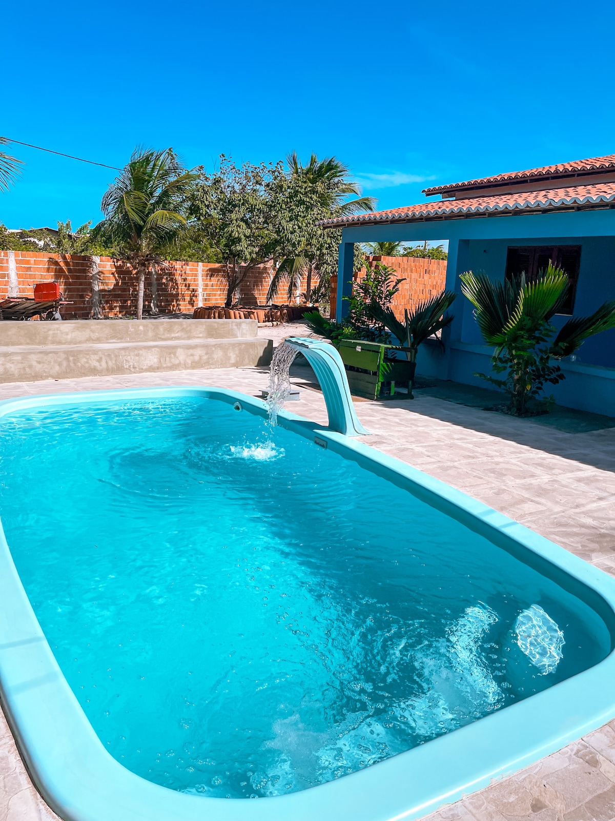 A rectangular swimming pool is set in an outdoor space, featuring a cascading water feature. Sunlight reflects off the clear blue water, surrounded by a stone patio and tropical plants. A shaded area with seating is visible in the background.