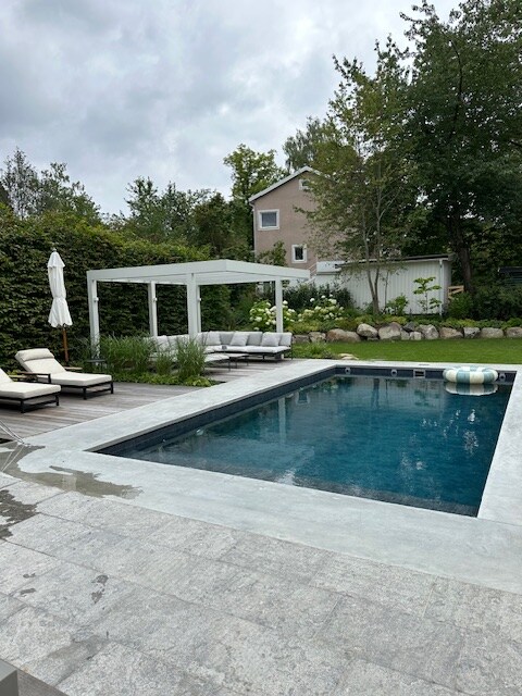 A rectangular outdoor pool is surrounded by stone paving and greenery, featuring two lounge chairs with cushions nearby. A shaded seating area offers protection from the elements, while a large inflatable ring rests on the pool's edge, inviting relaxation and leisure.