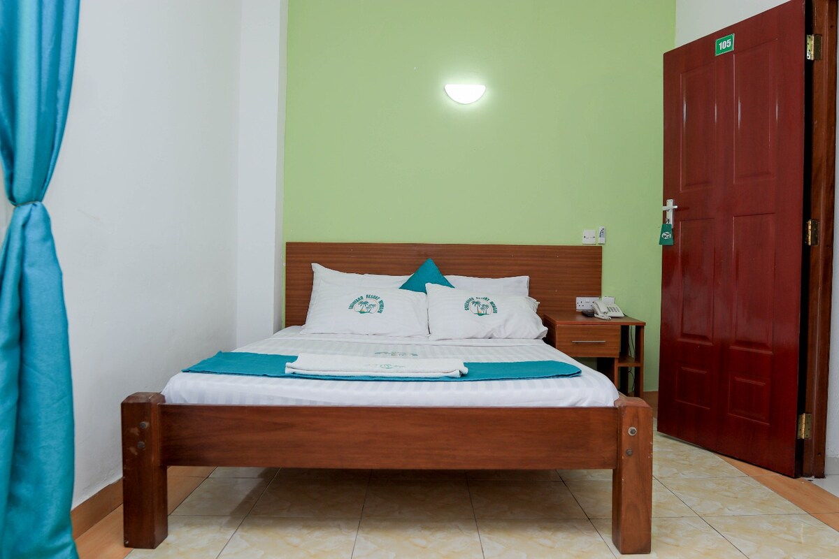 A cozy double room features a wooden bed with a padded headboard and crisp linens. The light green walls complement the turquoise accents, while a closed door is visible on the right. Soft, natural light is provided by a wall-mounted light fixture.