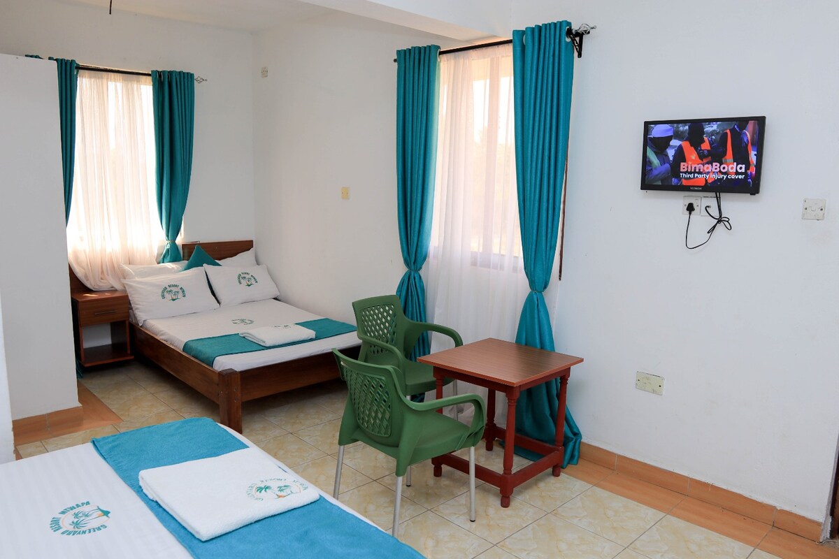 A room is shown with three single beds and soft turquoise curtains framing the windows. A small table and green chairs are placed in front of a wall-mounted television. Natural light fills the space, enhancing the inviting and functional design.