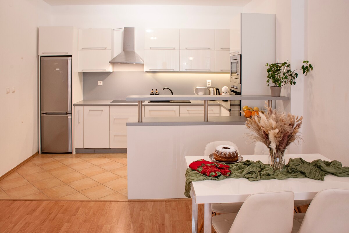 A modern kitchen is equipped with sleek, white cabinetry and stainless steel appliances, including a refrigerator, oven, and microwave. A dining area features a simple white table with four sleek chairs and a centerpiece of dried flowers. Natural light brightens the space.