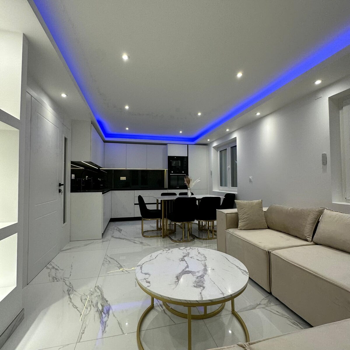 A modern living area features a light-colored sofa arranged around a marble coffee table. A sleek dining table set for six is visible, accompanied by stylish black chairs. Soft blue lighting enhances the contemporary design, while large windows allow natural light to enter.