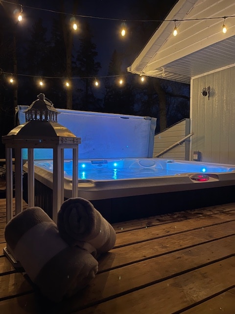 A hot tub is situated on a wooden deck, surrounded by twinkling string lights. Soft, fluffy towels are neatly placed nearby, and a lantern provides additional ambient lighting. The scene is illuminated with a calming blue glow from the hot tub.