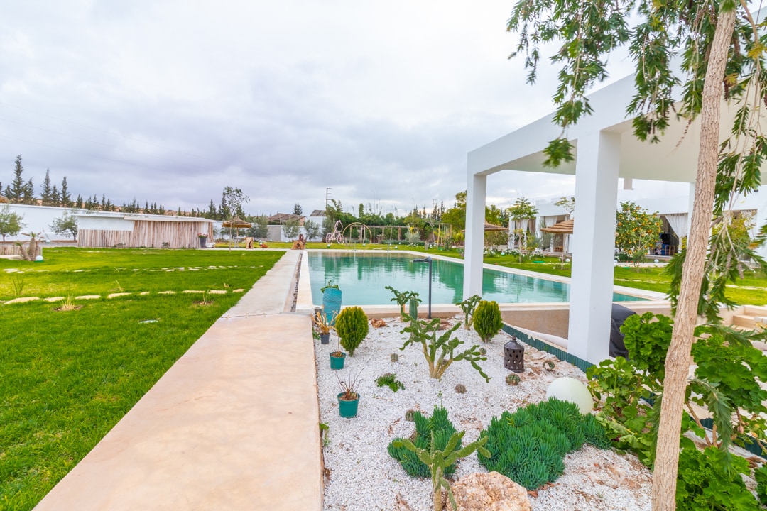 A spacious outdoor area is showcased, featuring a large pool surrounded by lush green grass and decorative plants. A covered seating area is visible beside the pool, providing shade. Softly landscaped paths lead into the gathering space, enhancing the serene atmosphere.