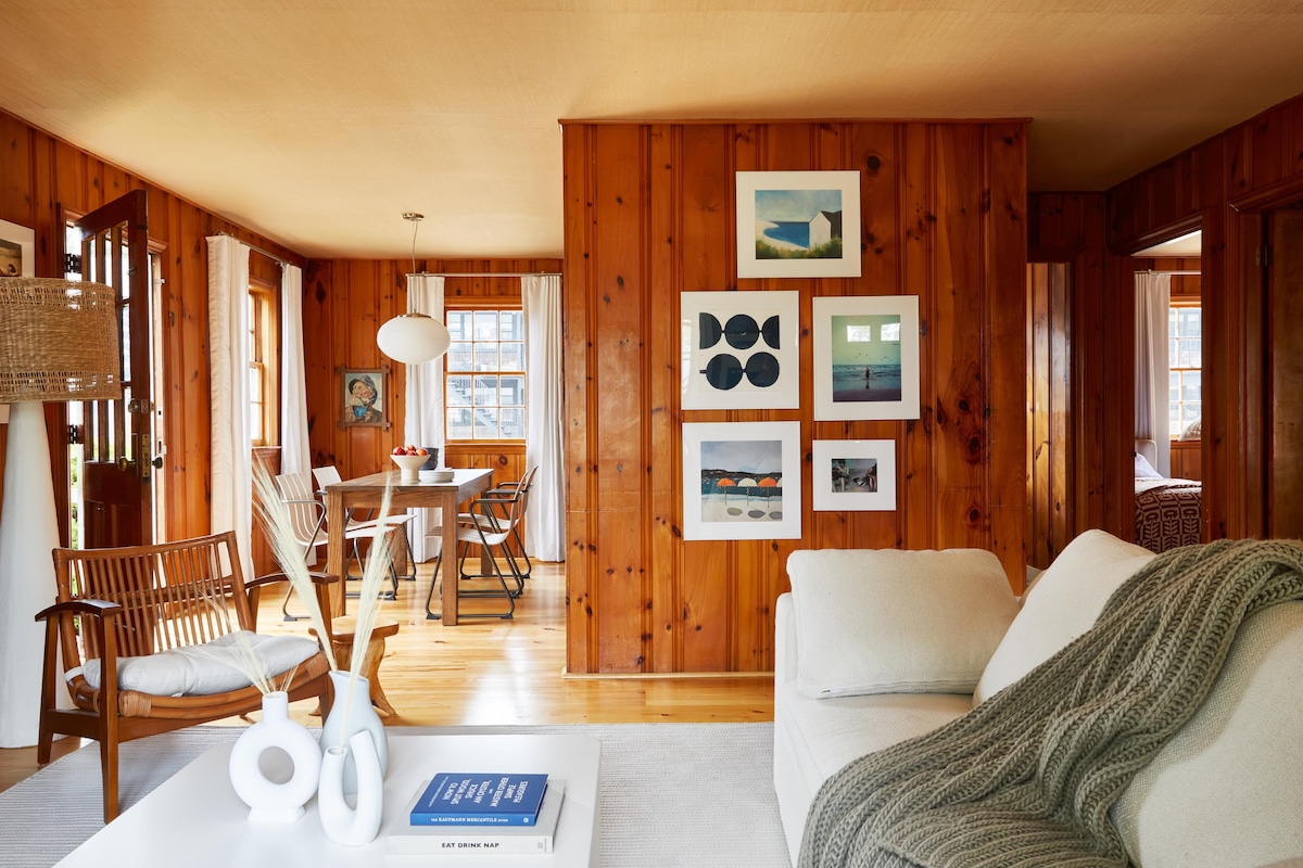 A cozy living area showcases wooden paneling, creating a warm environment. A comfortable couch is accompanied by a knitted throw. The space features a dining area with a round table and chairs, along with framed artwork displayed on the wall.