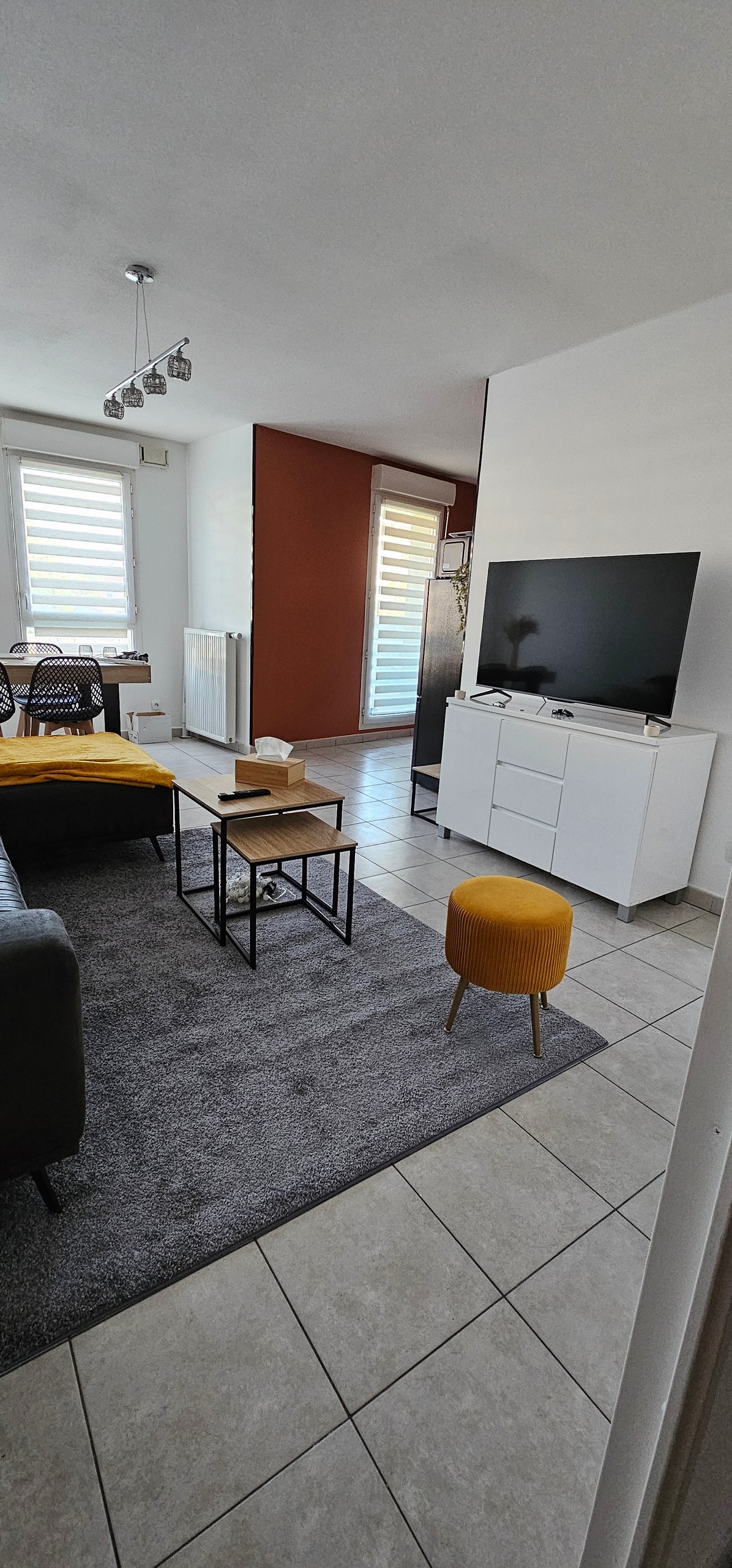 A modern living area features a comfortable couch and a stylish coffee table on a soft rug. A TV unit is positioned against the wall, with a large television and a bright yellow stool adding a pop of color. Natural light pours in through shaded windows.