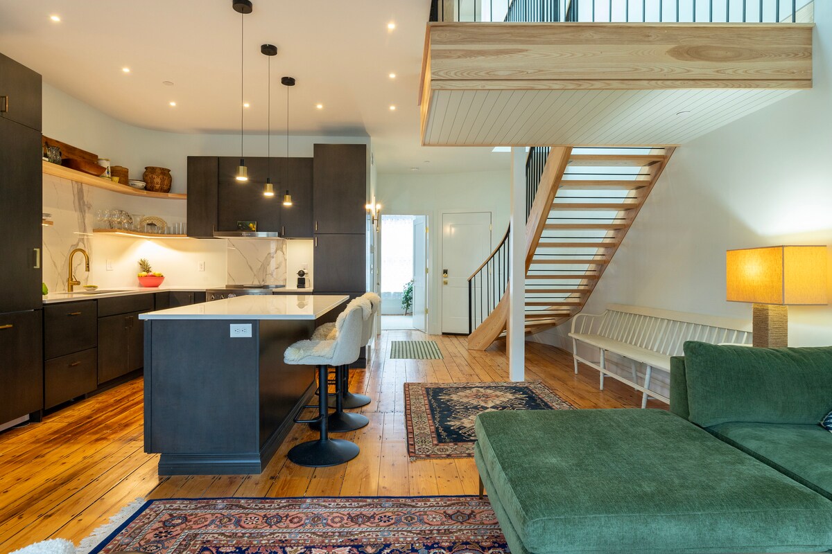 An open-plan living space showcases a modern kitchen with dark cabinetry and a white countertop. Bar stools are arranged at the kitchen island. A staircase with wooden steps leads to the upper floor. A green sofa sits adjacent to a rug, adding a touch of warmth.
