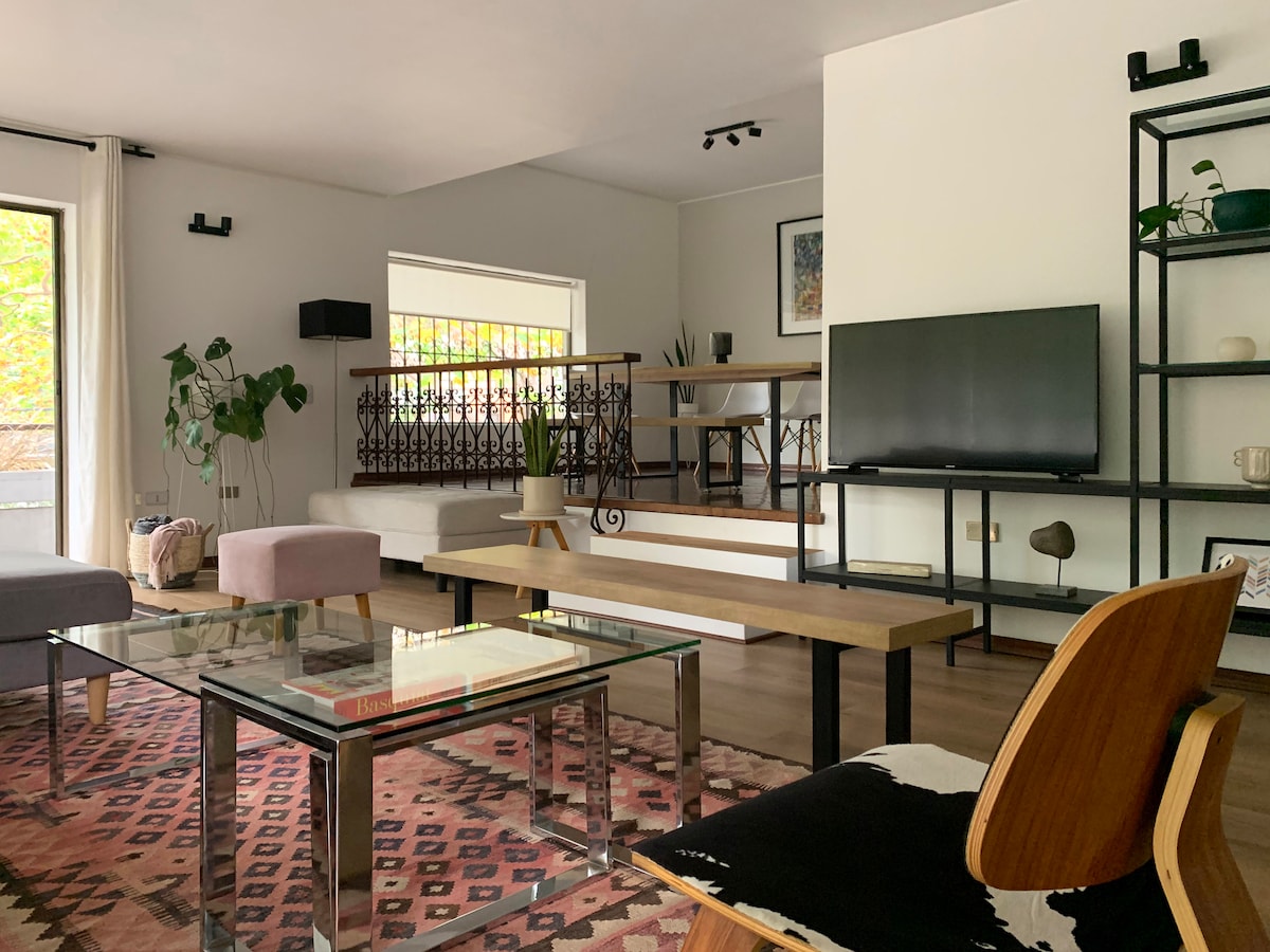 A spacious living area is presented with a combination of modern furniture and decorative elements. A large glass coffee table sits atop a patterned rug, with plush seating arranged nearby. Natural light enters through the large windows, enhancing the inviting ambiance of the room.