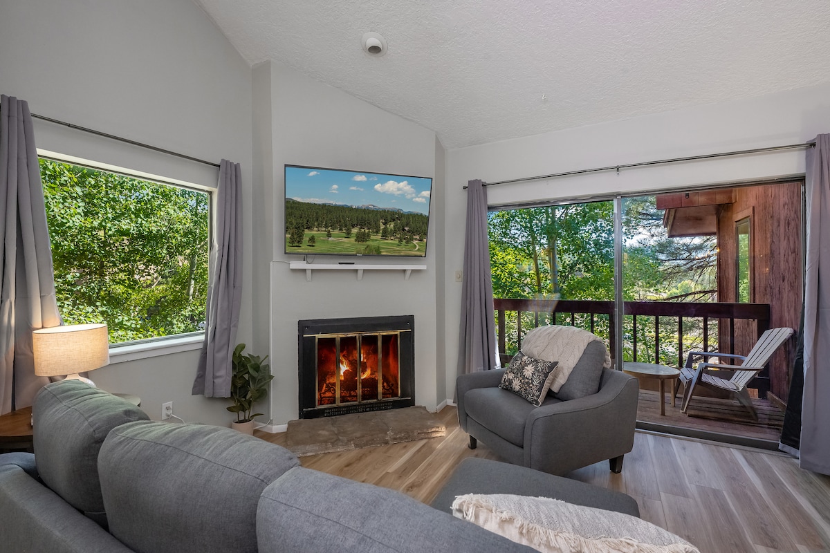 A cozy living area features a comfortable gray sofa and armchair, complemented by light curtains framing windows with views of greenery. A television is mounted above an electric fireplace, while a balcony with seating is visible through sliding glass doors.