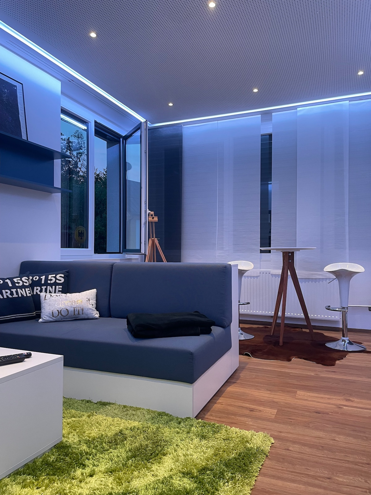 A modern living space features a sectional sofa against a light-colored wall, with vibrant green carpeting underfoot. Natural light enters through large windows adorned with sheer curtains. A high table with two stools is positioned nearby, complemented by a sleek floor lamp.