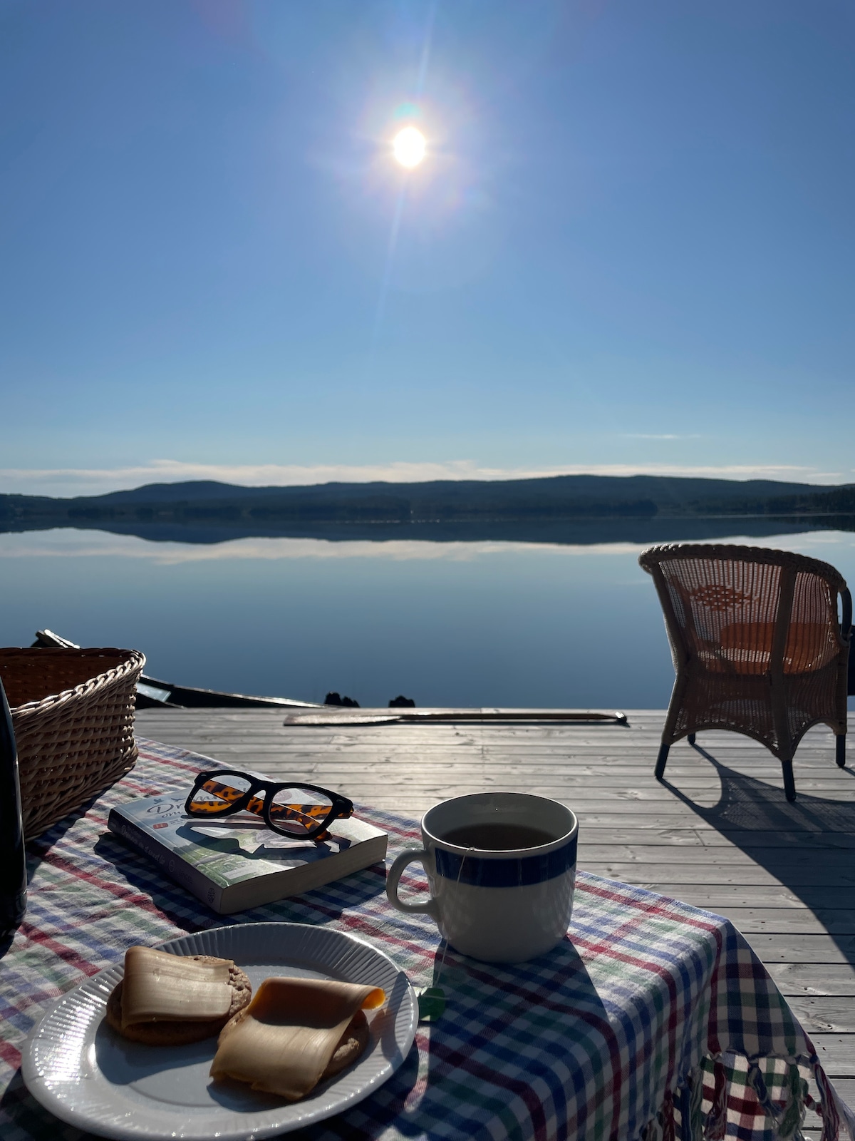 A serene lakeside scene captures a table set with a plate of snacks and a cup of coffee, accompanied by a book. A pair of sunglasses rests nearby, while a wicker chair offers a comfortable spot. The sun shines brightly in a clear blue sky, reflecting on calm waters.