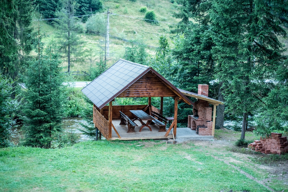 An outdoor gazebo is presented with a wooden structure and a sloped roof. A long table and benches are visible, offering seating for several guests. A stone barbecue is positioned nearby, surrounded by trees and a lush green lawn.