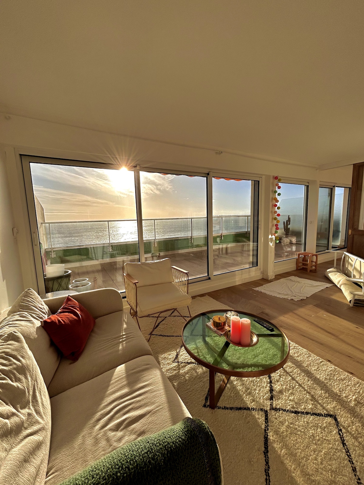 The living area features large glass sliding doors that open to a panoramic sea view. A beige sofa with a red accent pillow faces a round coffee table with candles. Natural light floods the space, illuminating a neutral area rug and an outdoor terrace visible in the background.