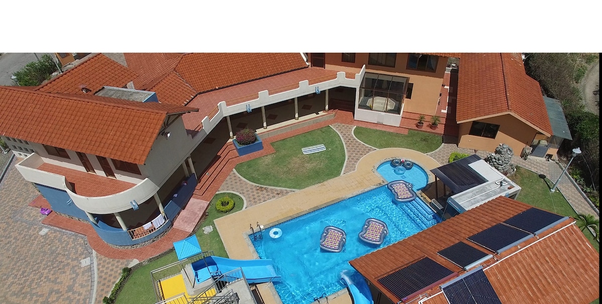 An aerial view showcases a spacious outdoor area featuring a swimming pool with loungers and jacuzzi sections. Surrounding the pool, pathways lead to various lounging areas, with the building's red-tiled roofs and green landscaping enhancing the environment.