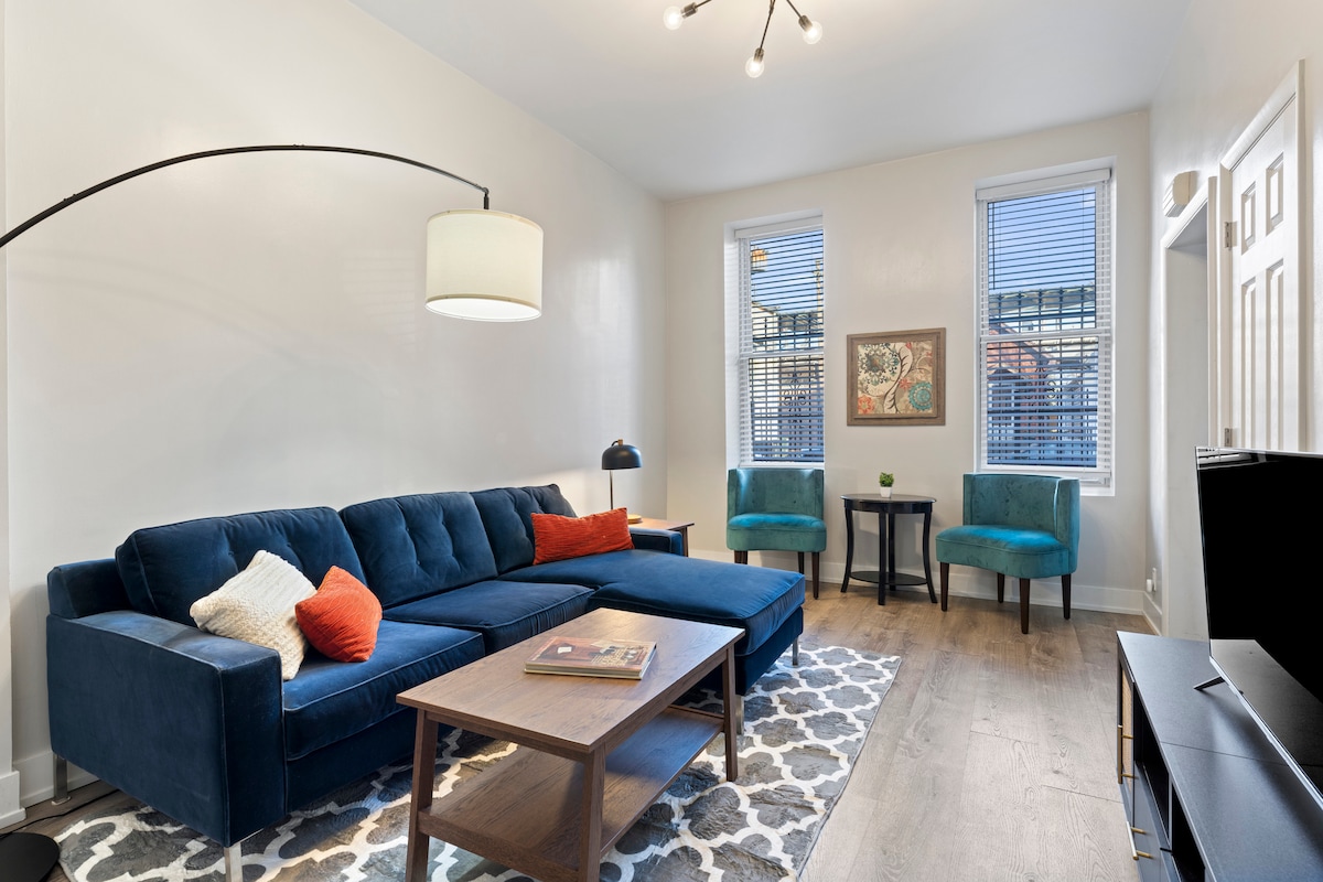 A spacious sitting room features a plush blue sectional couch with orange accent pillows, positioned across from a wooden coffee table. Two teal chairs complement the space, while framed artwork adds a touch of local flair. Natural light filters in through two windows adorned with blinds.