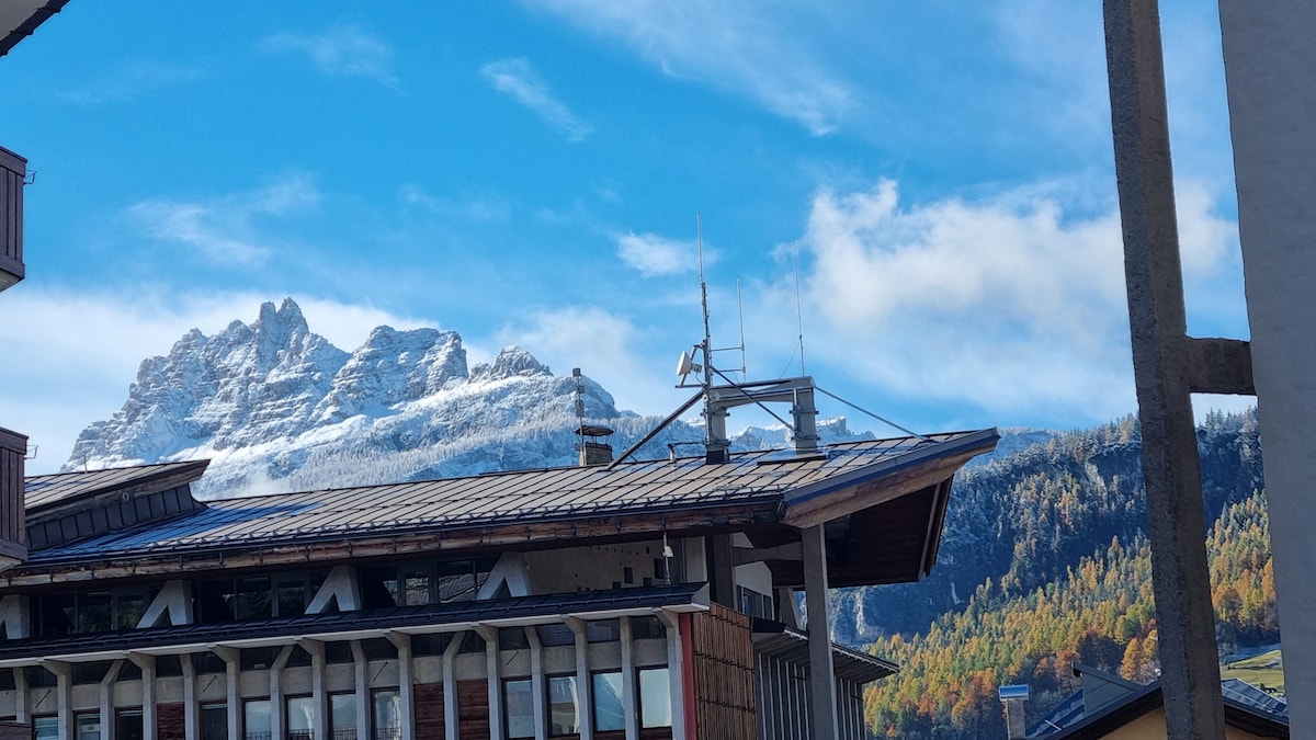 The image captures a view of the snow-capped mountains against a bright blue sky. Buildings are visible in the foreground, showcasing a mix of architectural styles. The vibrant colors of the trees in the valley indicate a scenic landscape typical of the region.