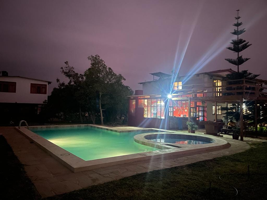 An illuminated outdoor pool area is visible, featuring a large rectangular pool and a smaller circular jacuzzi. Surrounding greenery and a modern structure with large windows create a serene atmosphere, while soft lighting highlights the space against a twilight sky.