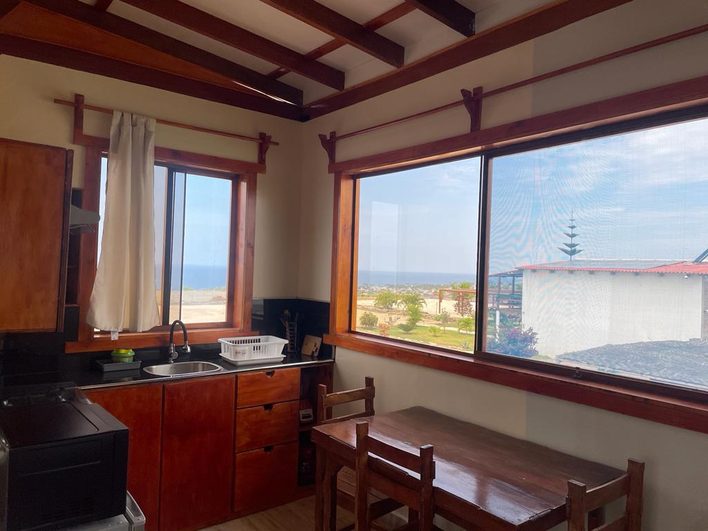 A bright kitchen area features wooden cabinets and a sink beside a large window offering views of the outdoor landscape. A wooden dining table with chairs is positioned near the window, allowing for comfortable dining while enjoying the sight of the ocean in the distance.