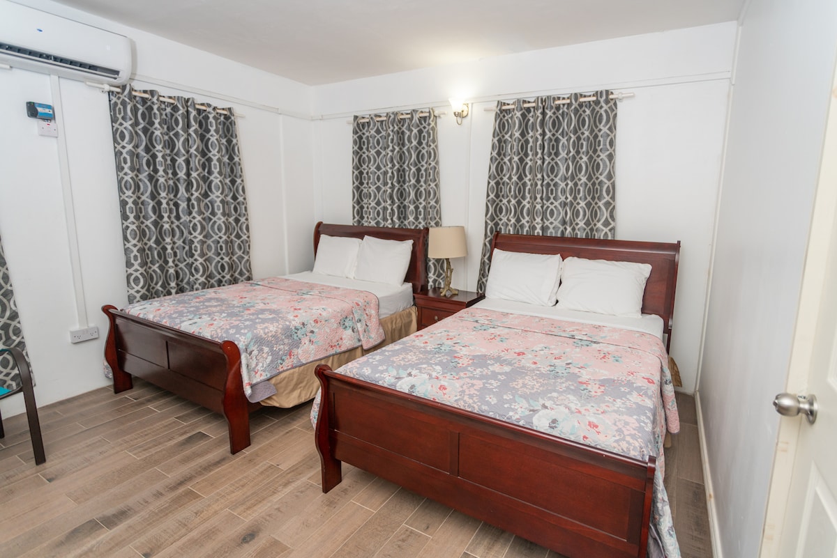 A well-furnished room features two spacious beds with floral-patterned bedding. Light-colored curtains frame the windows, allowing natural light to partially enter. A bedside lamp is positioned between the beds, and a subtle wooden floor adds warmth to the space.