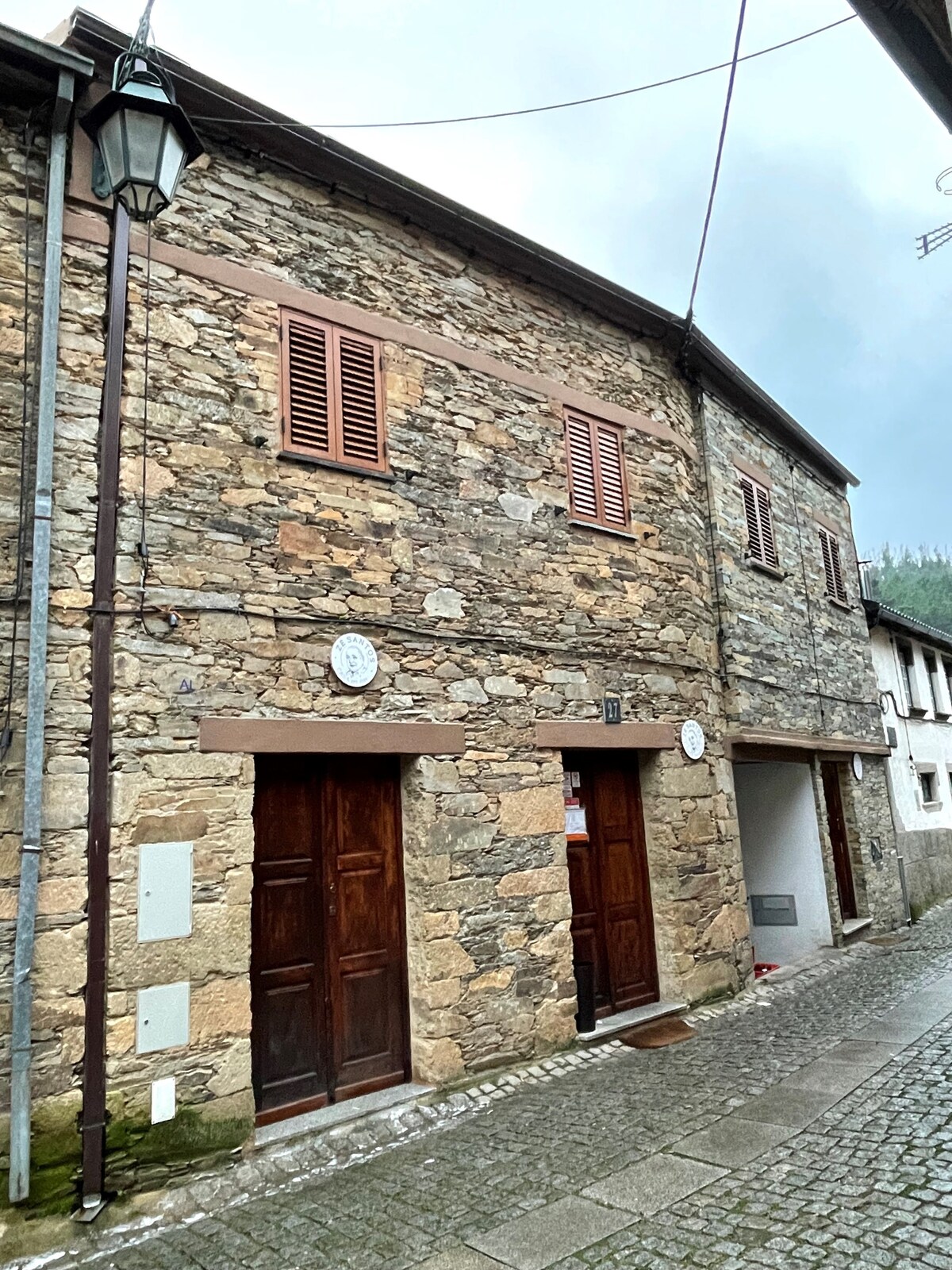 A rustic stone building with wooden doors stands along a cobblestone street. The exterior features shuttered windows and decorative circular elements. A street lamp is visible, illuminating the area. The surrounding environment includes other traditional structures, creating a charming village atmosphere.