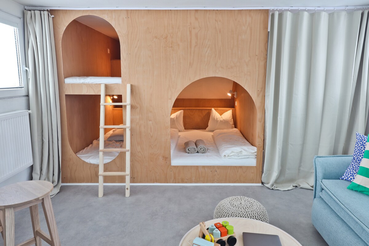 The interior features a wooden wall with two cozy sleeping nooks, each equipped with soft bedding and individual lighting. A compact ladder offers access to the upper sleeping area. A comfortable seating area is present, complemented by a small table with playful items.