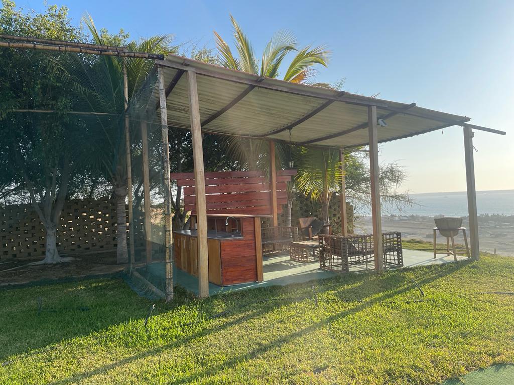 An open-air seating area features natural wood furniture under a shaded roof. Lush green grass surrounds the space, providing a comfortable outdoor environment. In the background, a scenic view of the horizon and a glimmering body of water are visible, offering a serene atmosphere.