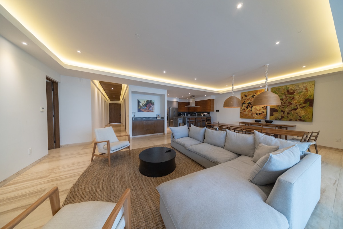 A spacious living room showcases a large sectional sofa, ivory walls, and recessed lighting. Natural textures are present with a woven area rug and wooden accents. A dining area, visible in the background, features a long table with multiple chairs. Artwork adorns one corner, enhancing the environment.