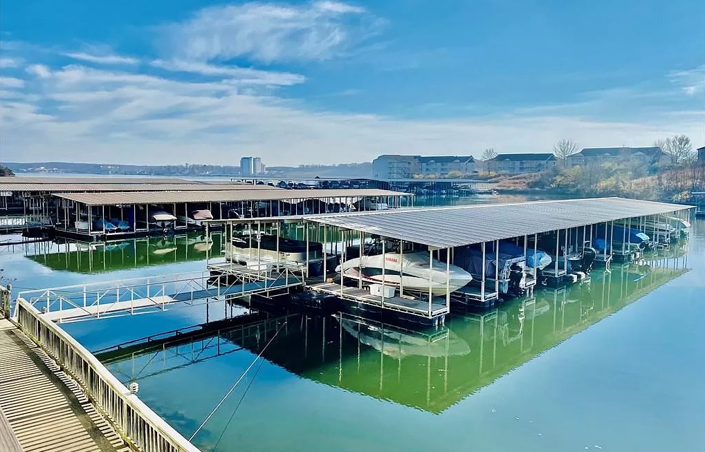 A community dock is visible with numerous boat slips outlined against the calm water, reflecting the clear blue sky. The area showcases sheltered spaces for multiple boats, creating a serene lakeside environment.