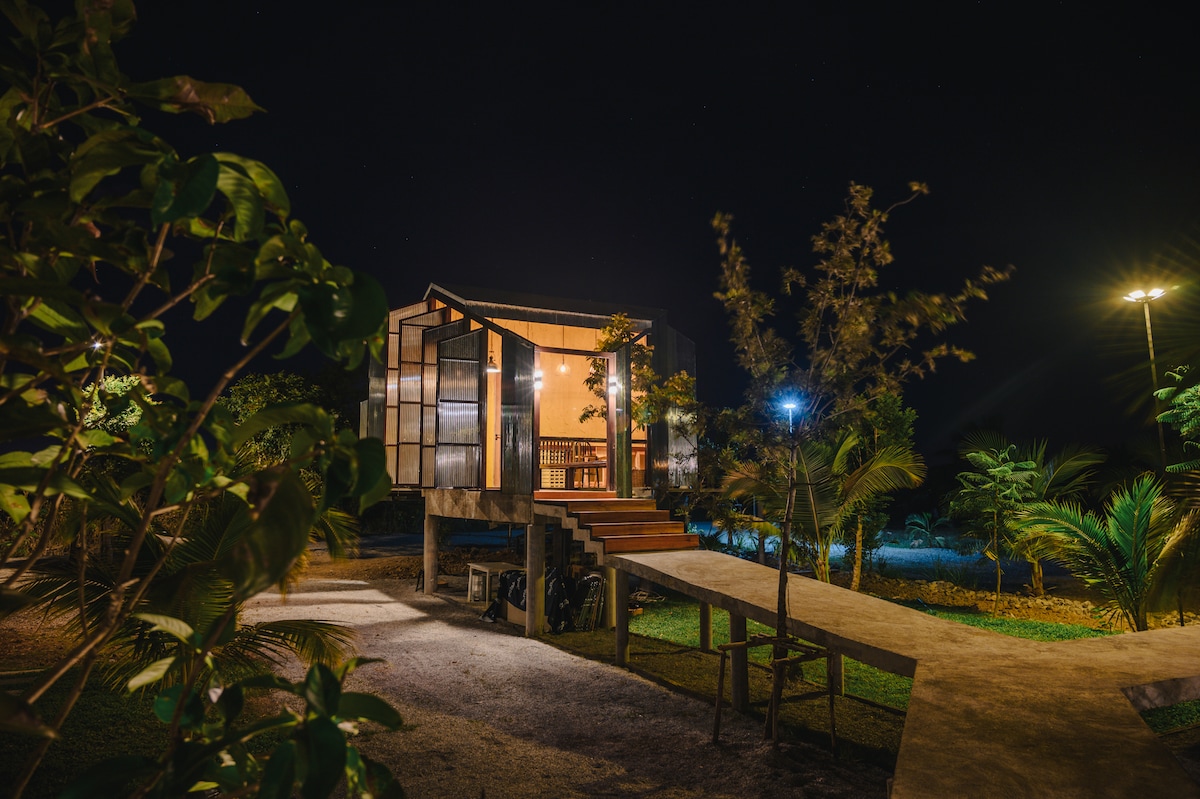 The cabin is illuminated at night, showcasing its unique architecture with large glass windows. Surrounding greenery and pathway lights create a serene atmosphere, while the elevated structure is accessed by a set of stairs leading to a spacious deck.