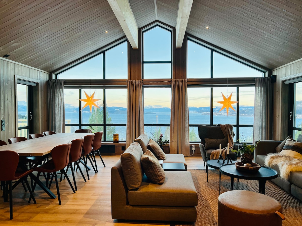 A spacious living area features large windows providing panoramic views of the fjord. A comfortable seating arrangement includes a sofa with decorative cushions. A dining table with modern chairs sits nearby. Natural light fills the room, complemented by minimalist decor and soft textures.