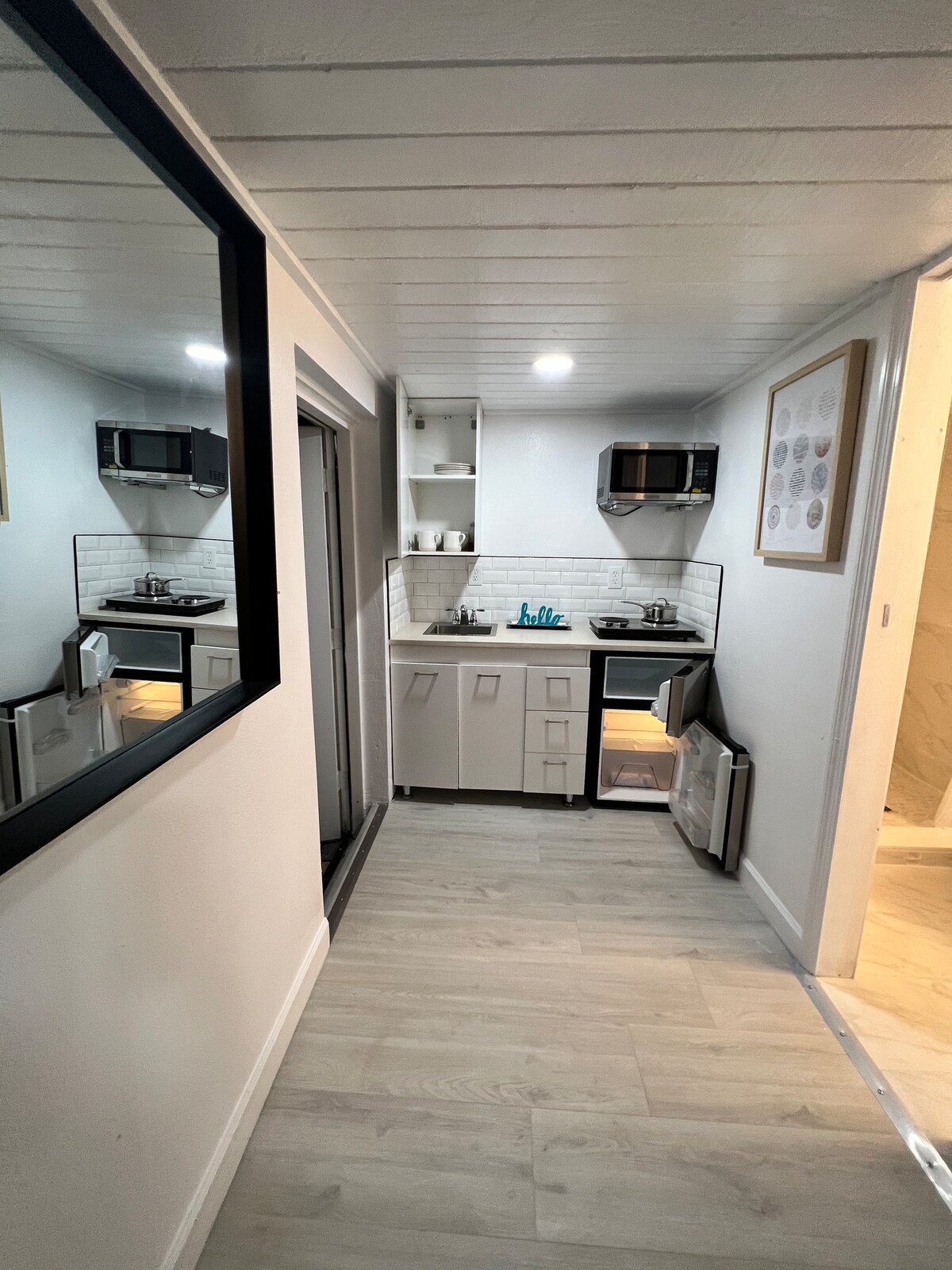 A compact kitchen area is visible, featuring dual stovetops, a large sink, and a refrigerator. Modern cabinetry provides storage space, while light fixtures illuminate the countertop. A mirror on the left reflects part of the space, enhancing its brightness and depth.