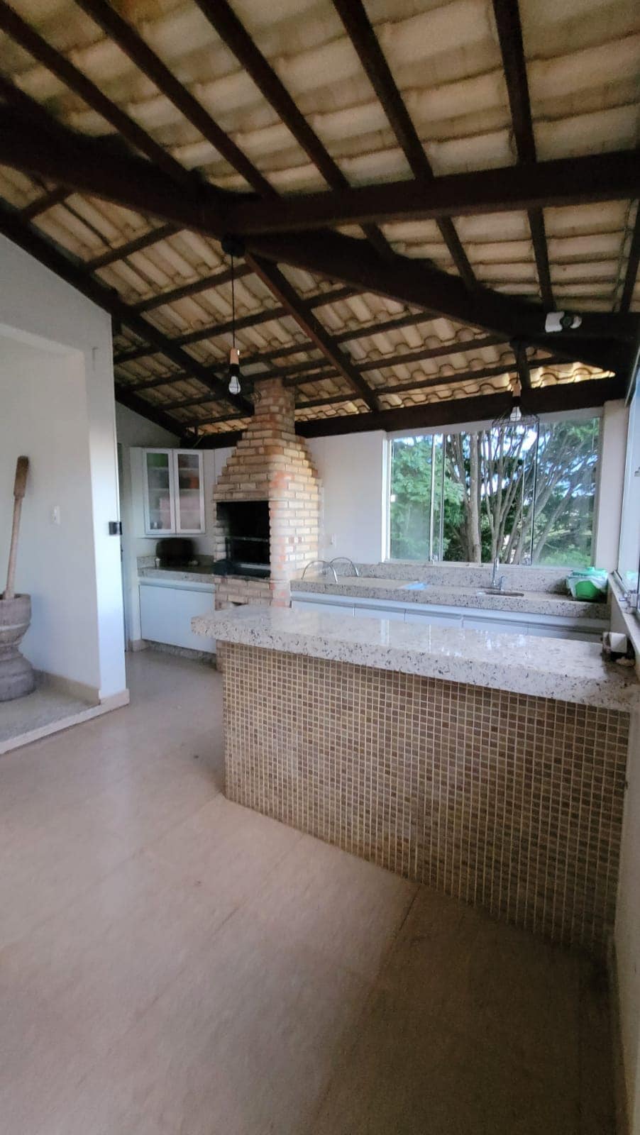 The kitchen area features a high, open ceiling with wooden beams, allowing natural light to enhance the space. A brick fireplace is set against one wall. The kitchen counter is made of stone, bordered by tiles that add a textured element.