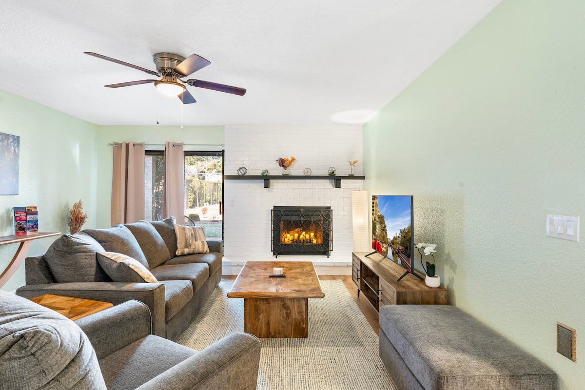 A spacious living area features a comfortable sectional sofa and armchair arrangement around a wooden coffee table. A fireplace adds a cozy touch, while a television is positioned on a media console. Large windows allow natural light to fill the room, complemented by light green walls.