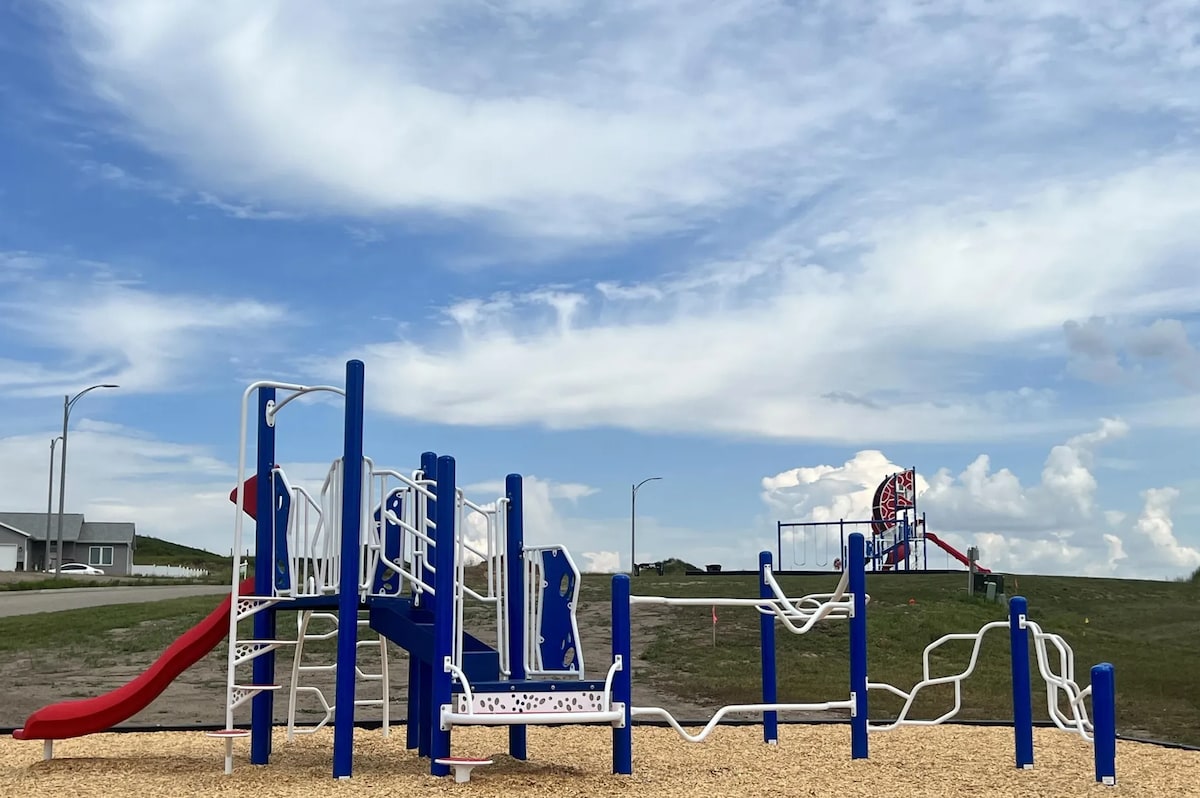 A modern playground is displayed, featuring brightly colored equipment, including a slide and climbing structures. Play areas covered with soft mulch are visible, with blue, white, and red colors providing a playful aesthetic. Open grassy areas surround the playground, under a blue sky with fluffy white clouds.