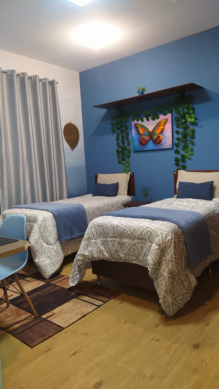 A bright blue room features two twin beds with patterned bedding. A nature-themed wall art piece is displayed above a small plant on a shelf. Soft curtains frame a window, enhancing the serene atmosphere, while a desk and chair are positioned in the corner.