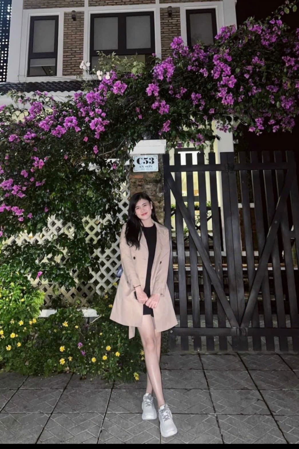 A charming entrance is framed by vibrant purple bougainvillea, cascading over a wooden gate. The paved pathway leads to a two-story building with large windows, providing a glimpse of the inviting interior behind the gate.