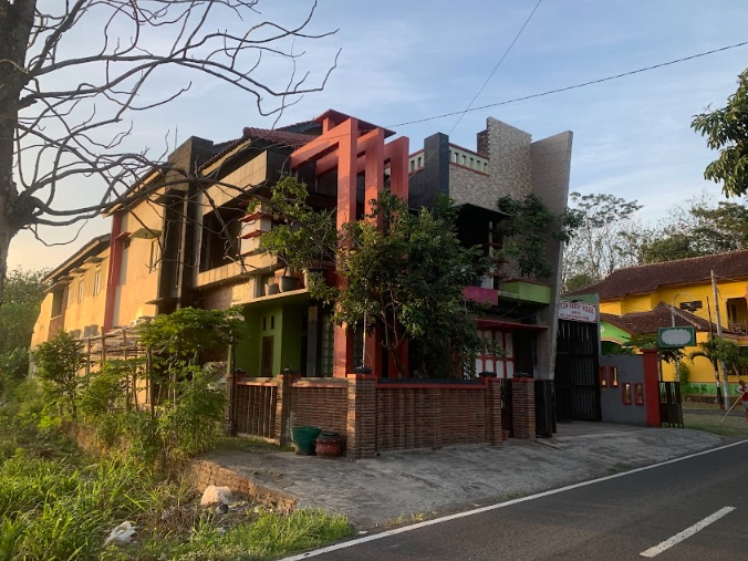 A unique multi-story building features a combination of vibrant colors and textured surfaces. Green foliage surrounds the structure, while a paved path leads to the entrance. The architecture demonstrates a blend of modern and traditional styles, providing an eye-catching exterior.