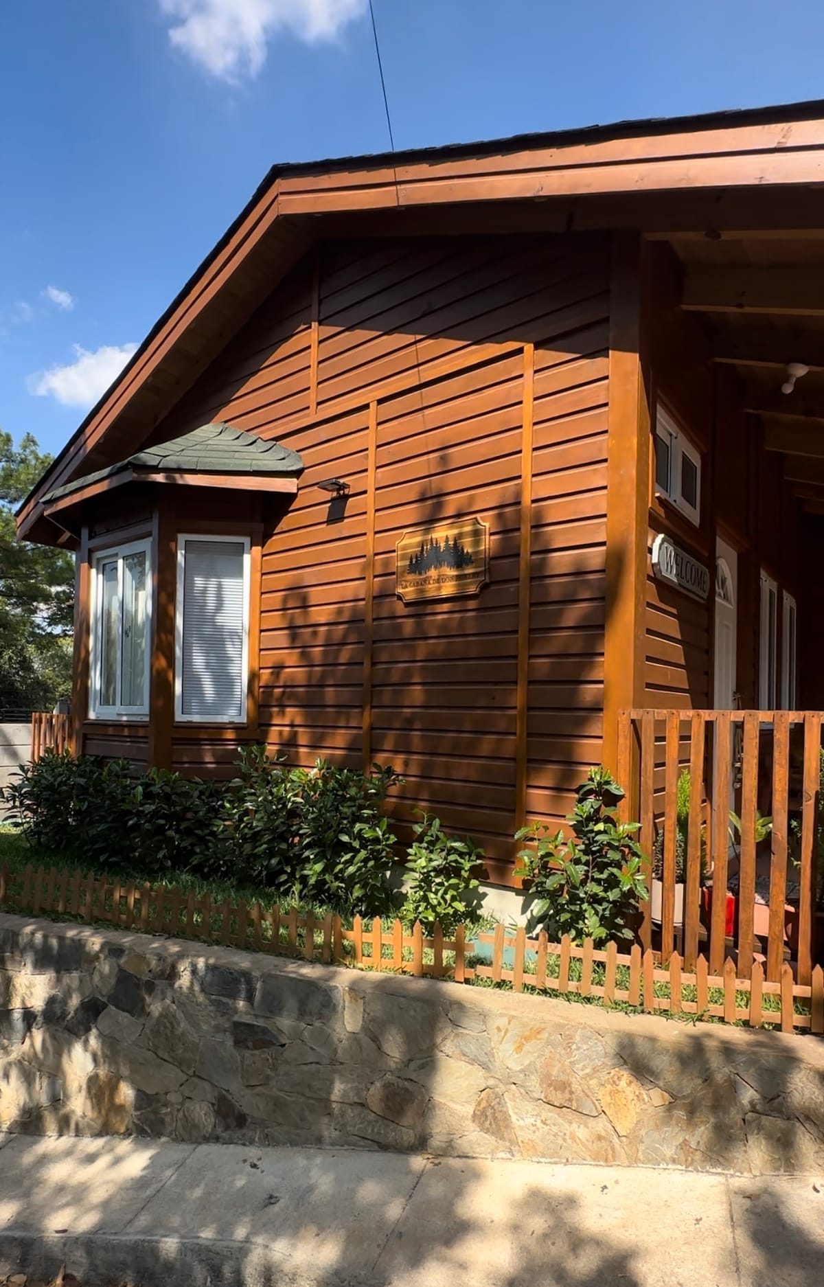 The exterior of a rustic wooden cabin is displayed, showcasing natural wood paneling and large windows. A low stone wall lines the front, complemented by a wooden fence and lush greenery, offering a warm and inviting entry to the space.