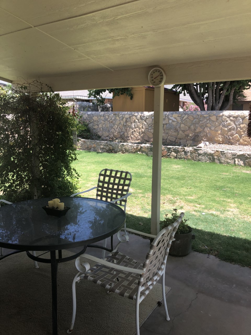 A shaded outdoor seating area features a round glass table surrounded by four white metal chairs. A lush grass backyard is visible, framed by a textured stone wall and greenery, providing a peaceful environment for relaxation.