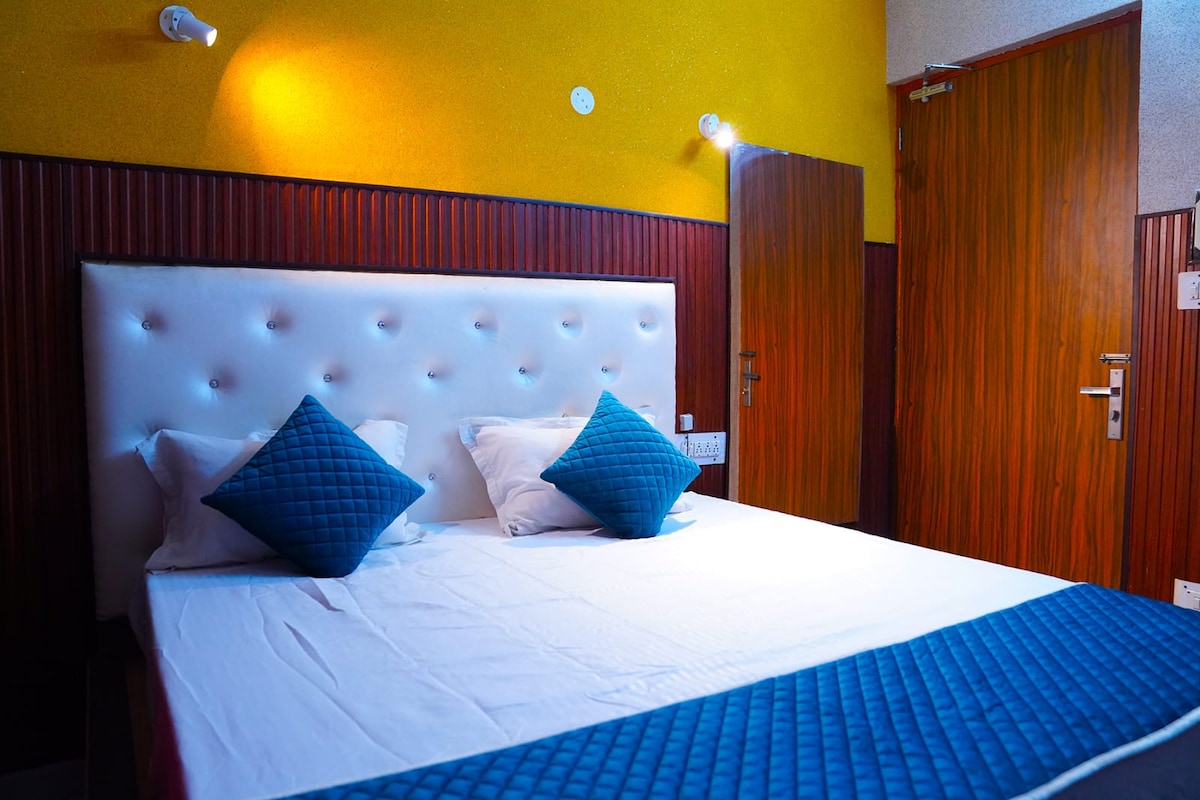 A comfortable bedroom features a headboard with a tufted design and two decorative blue cushions. Light from wall-mounted fixtures brightens the textured yellow and brown walls, while a wooden door provides access to the room.