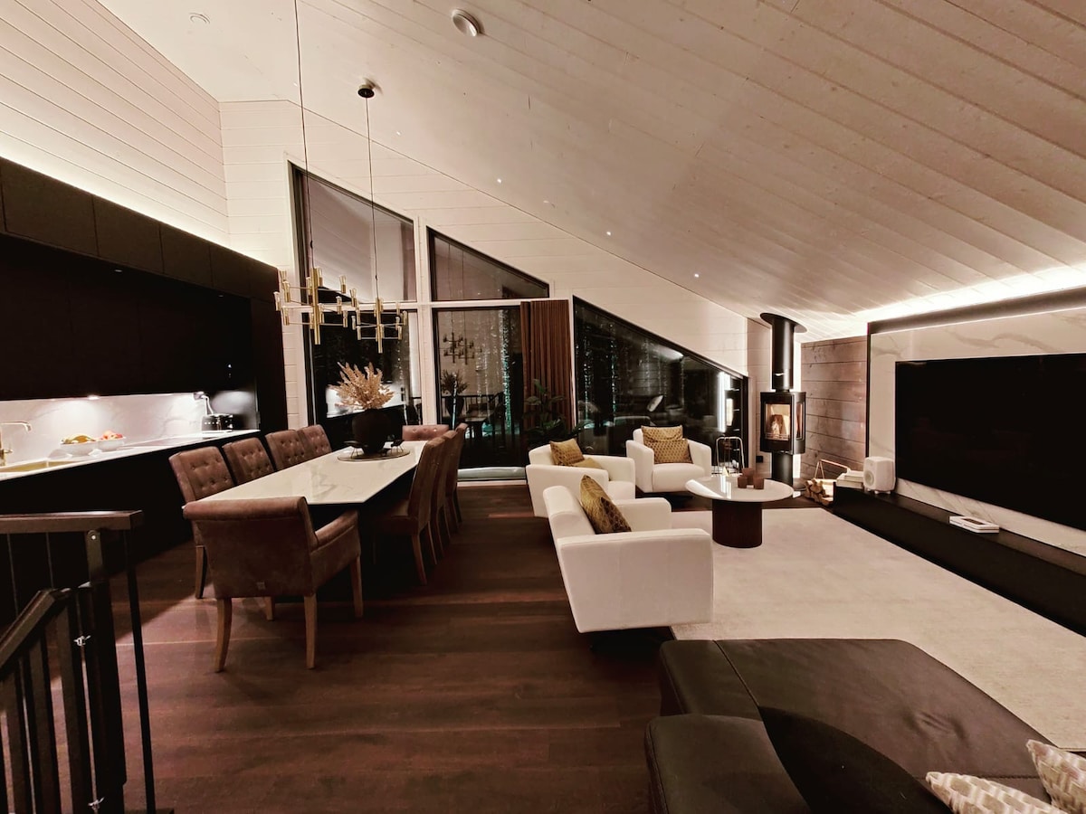 An open living space features high ceilings with wooden paneling, enhancing the airy feel. A dining table with upholstered chairs is visible next to a modern kitchen area. A comfortable seating arrangement surrounds a coffee table, with a stylish fireplace and large windows providing natural light.
