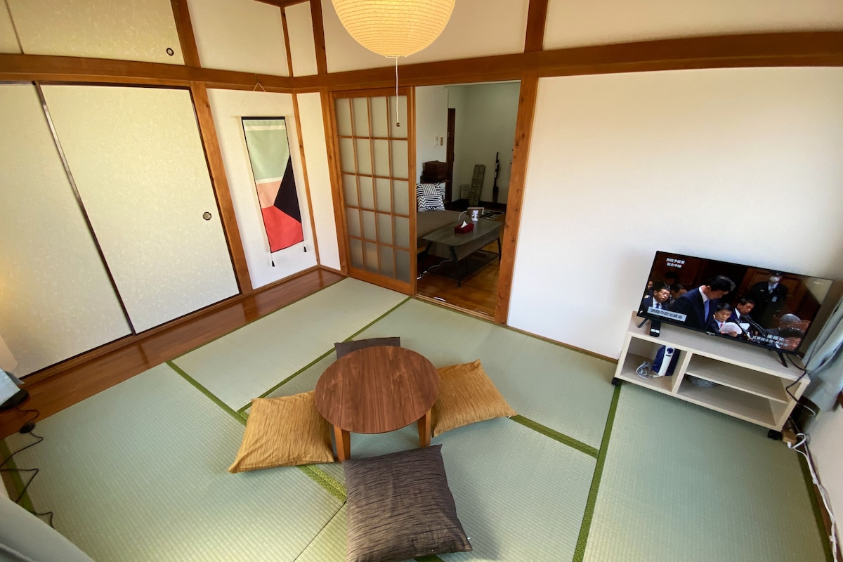 A traditional room with tatami flooring is shown, featuring a low round table at the center surrounded by floor cushions. A television is positioned on a stand, and shoji screens are visible in the background, enhancing the room's serene atmosphere.