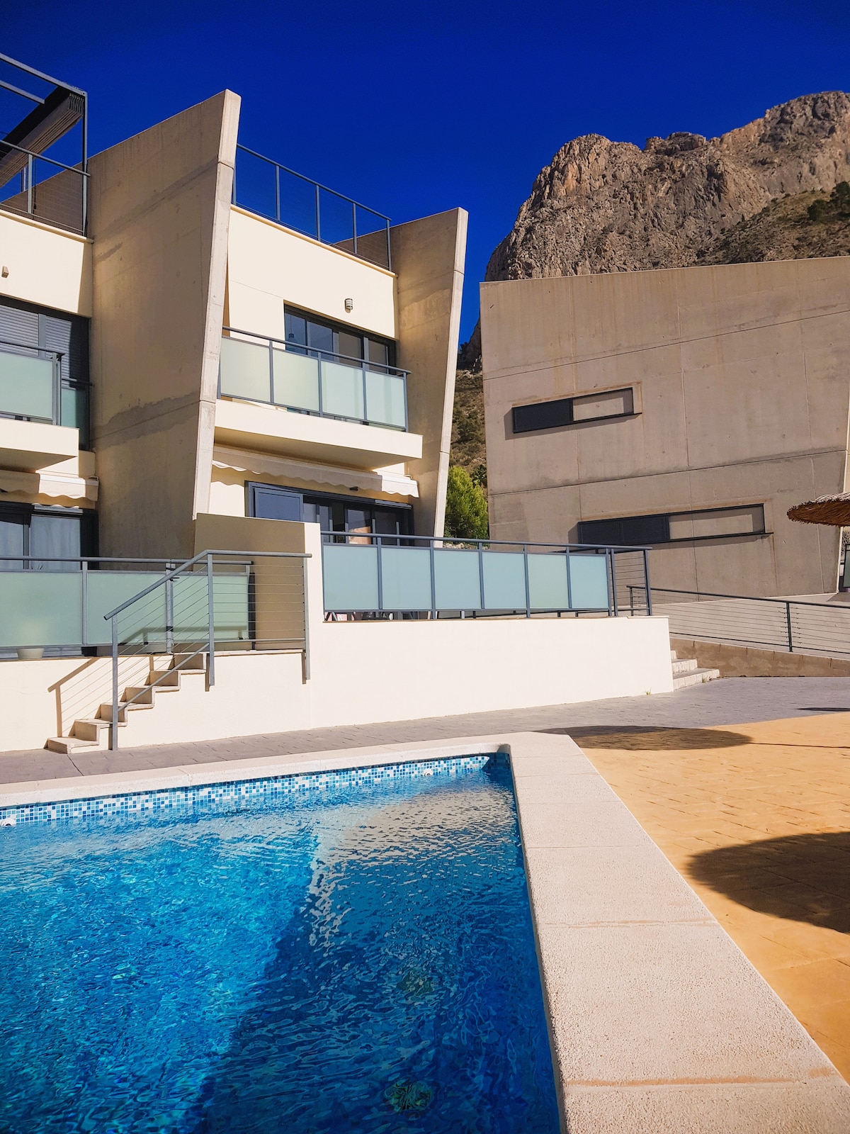 A modern building is seen alongside a swimming pool framed by a textured deck. Clear glass railings provide a view of the mountain backdrop, complemented by bright blue skies. The design features multiple levels with balconies, enhancing the contemporary aesthetic.