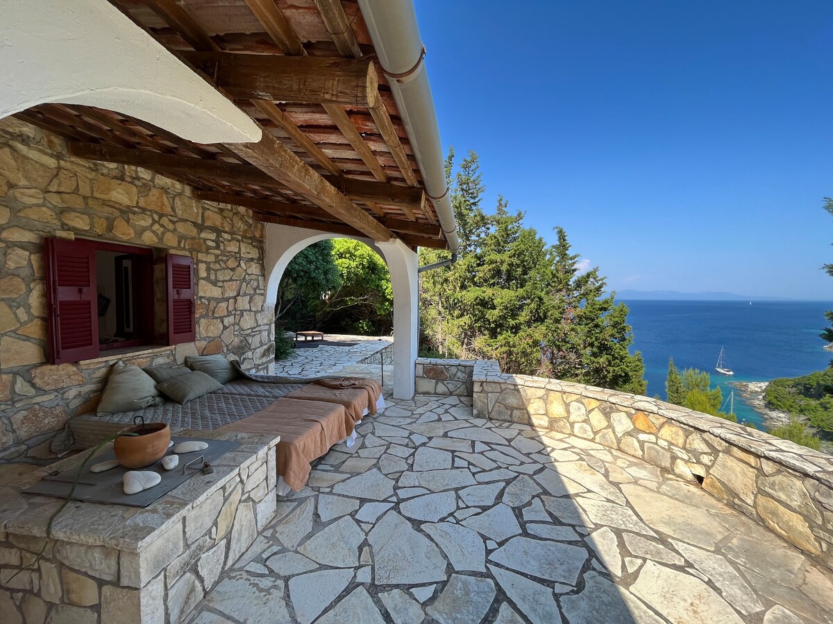 A stone terrace is presented, featuring a relaxed seating area with plush cushions and a textured blanket. Lush greenery surrounds the space, and a calm sea is visible in the background, with sailboats gently bobbing on the water under a clear blue sky.